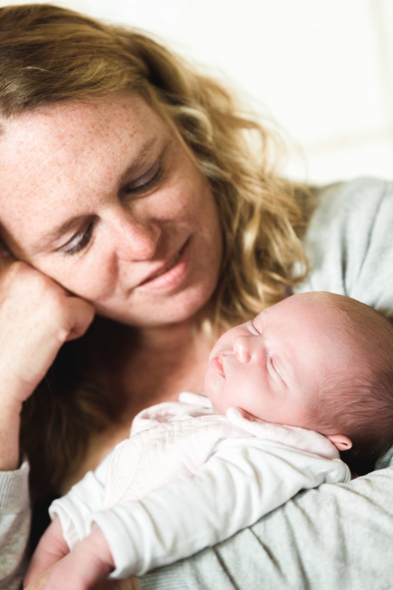 Une femme aux cheveux blonds ondulés sourit doucement et regarde un nouveau-né endormi bercé dans ses bras. Le bébé, vêtu d'une tenue blanche, semble paisible et satisfait. La femme repose sa tête sur sa main, dégageant une expression sereine et aimante dans ce moment de naissance sereine.