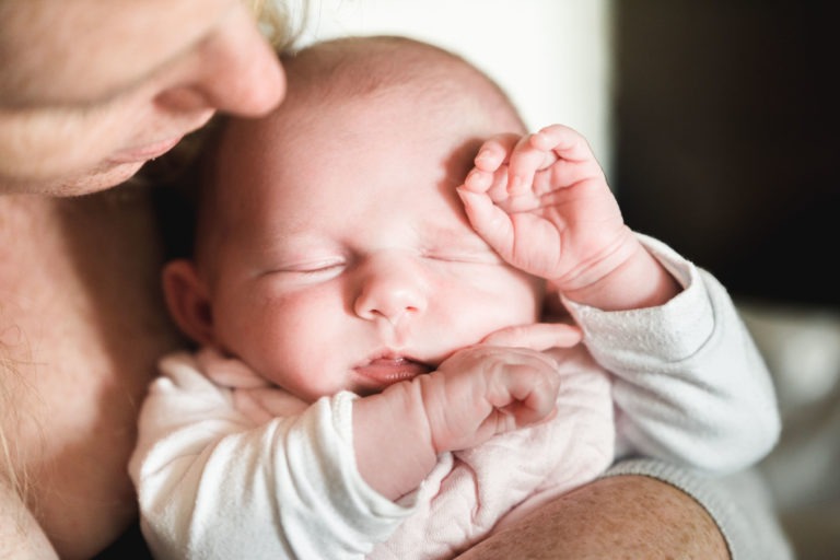 Gros plan d'un bébé endormi reposant dans les bras d'un adulte. Le bébé, incarnant une naissance sereine, porte une tenue de couleur claire et a une main levée près du visage. L'adulte est partiellement visible, berçant étroitement le bébé, le visage tourné vers l'enfant, semblant affectueux et protecteur.