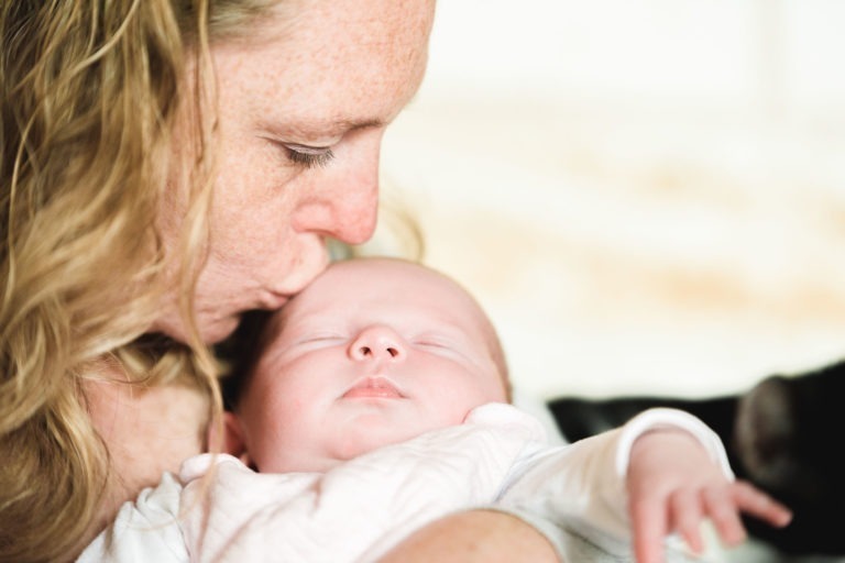 Gros plan d'une femme aux cheveux blonds ondulés embrassant doucement le front d'un bébé endormi. Le bébé, vêtu d'une tenue blanche et les yeux fermés, incarne Naissance sereine. Ce moment intime capture la paix et l'affection entre eux, sur fond légèrement flou.