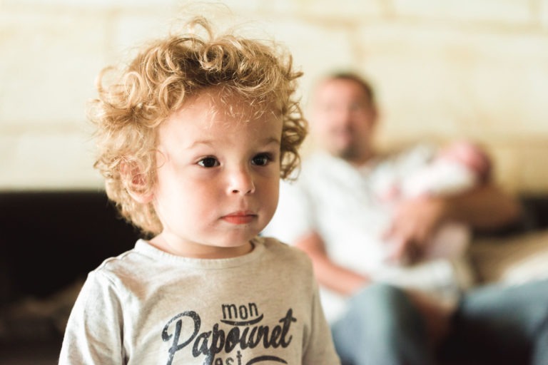 Un jeune enfant aux cheveux blonds bouclés regarde légèrement hors champ avec une expression sérieuse. L'enfant porte une chemise de couleur claire avec du texte dessus, bien que le texte soit partiellement obscurci. Dans l'arrière-plan flou, un adulte tient un bébé, évoquant une sensation de naissance sereine dans un cadre intérieur.