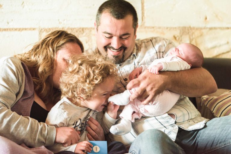 Une famille de quatre personnes est assise sur un canapé. La mère, aux cheveux châtain clair, câline un bambin aux cheveux bouclés qui tient un petit livre. Le père, barbu et moustachu, berce avec amour un nouveau-né dans ce moment de naissance sereine. Tout le monde apparaît heureux et engagé contre un mur de couleur claire.