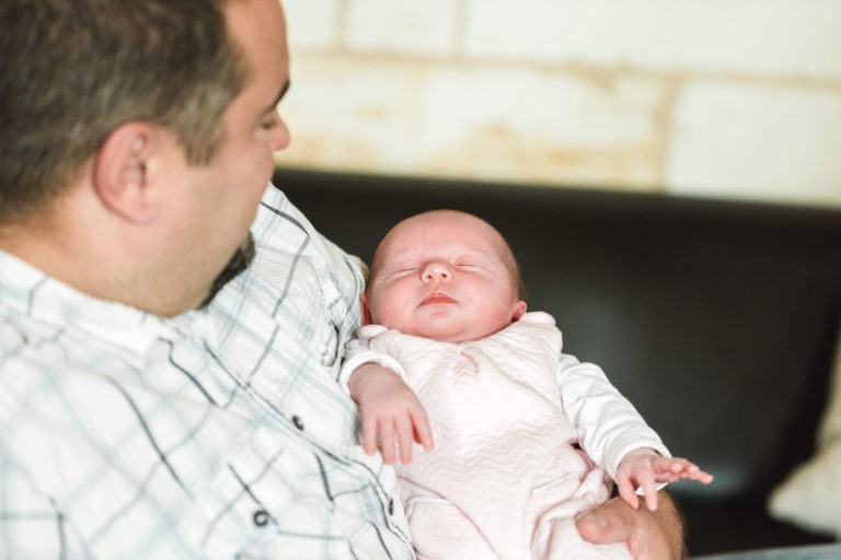 Un homme en chemise à carreaux tient dans ses bras un bébé endormi, vêtu d'une combinaison rose. Le bébé, incarnant Naissance sereine, repose paisiblement dans les bras de l'homme tandis qu'il regarde l'enfant avec amour. L'arrière-plan est flou, avec un mur de briques blanches et un canapé sombre.