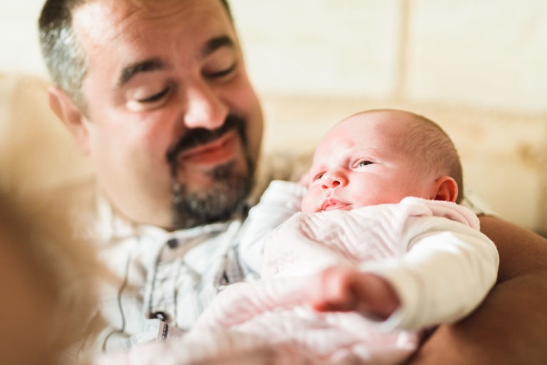 Un homme souriant aux cheveux courts et à la barbe berce un nouveau-né vêtu de blanc. Le bébé, qui regarde vers le haut dans une combinaison blanche avec une couverture à motifs autour, dégage un air de naissance sereine. L'arrière-plan est légèrement flou, mettant l'accent sur le moment tendre entre l'homme et le nourrisson.