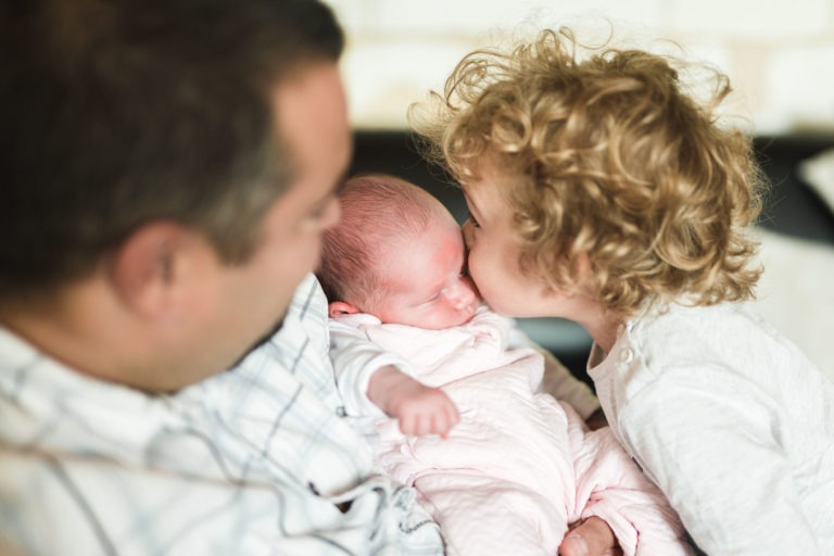 Un homme tient dans ses bras un nouveau-né emmailloté. Un enfant aux cheveux bouclés, vêtu de blanc, se penche pour embrasser le bébé sur le front. La scène capture un moment de tendresse familiale de naissance sereine, mettant l'accent sur l'affection et la chaleur partagées entre les membres de la famille. L'arrière-plan est légèrement flou.