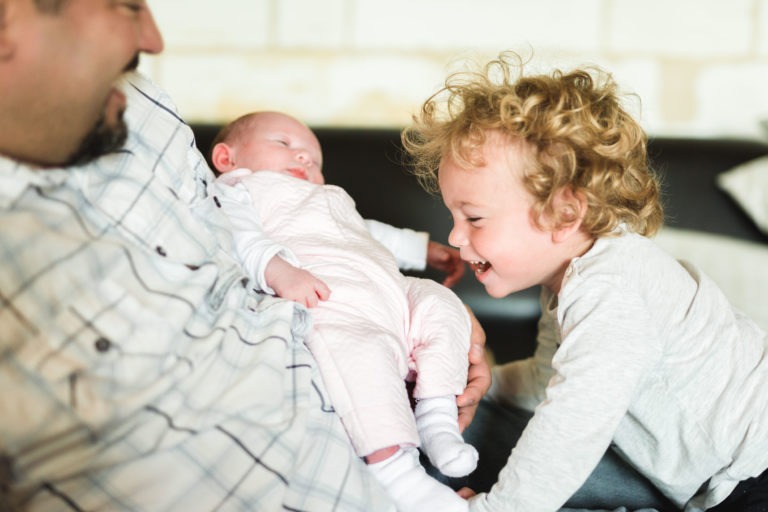 Un homme, partiellement visible, tient dans ses bras un bébé endormi, habillé de blanc. Un bambin aux cheveux bouclés à droite se penche vers le bébé avec une expression de joie. Ils sont à l'intérieur, dans un décor chaleureusement éclairé, suggérant un moment familial rempli d'affection et célébrant cette naissance sereine.