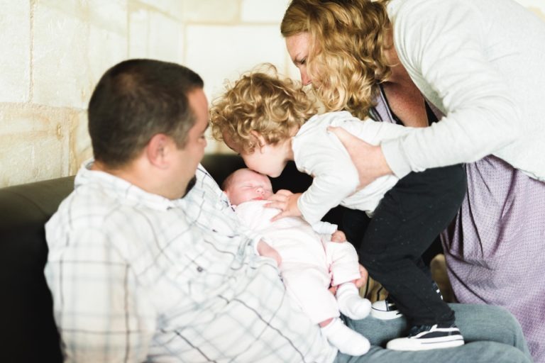 Un moment de tendresse en famille : un homme est assis sur un canapé, berçant un bébé habillé de blanc, tandis qu'une femme debout se penche pour aider un jeune enfant à embrasser la tête du bébé. Les membres de la famille, habillés de façon décontractée, sont dans une pièce chaleureusement éclairée aux murs clairs, célébrant cette naissance sereine.