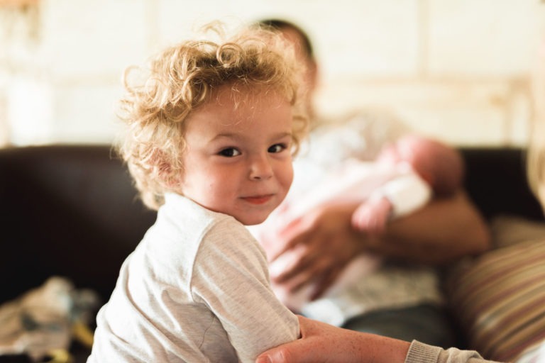 Un jeune enfant aux cheveux bouclés et au sourire espiègle regarde vers la caméra. En arrière-plan, un adulte berce un bébé, enveloppé dans une couverture rose pâle. La scène respire la sérénité, avec un éclairage doux et des tons doux, donnant un sentiment de proximité familiale et de bonheur.