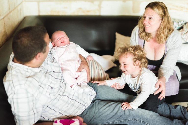 Une femme souriante aux cheveux blonds ondulés est assise sur un canapé à côté d'un homme en chemise à carreaux tenant un bébé qui pleure. Un bambin aux cheveux bouclés, vêtu d'une chemise à manches longues et d'un pantalon, grimpe joyeusement sur la jambe de l'homme. La famille respire la sérénité tandis qu'ils se détendent dans leur cadre douillet aux murs clairs et au canapé sombre.