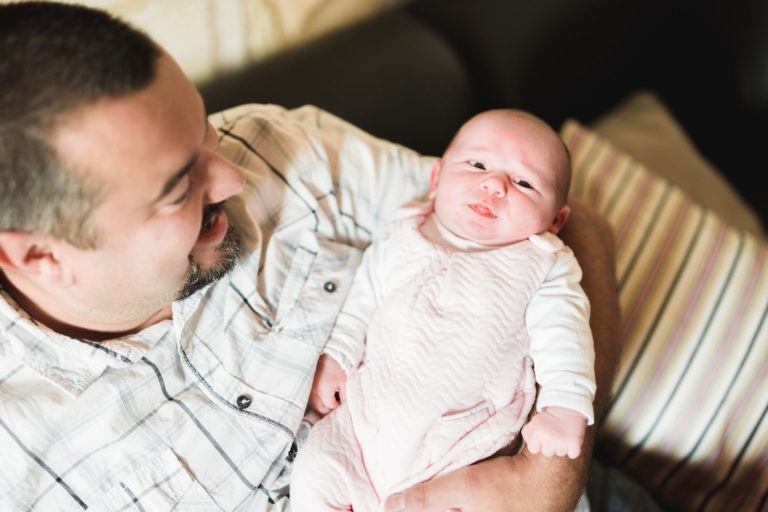 Un homme aux cheveux noirs courts et à la barbe, vêtu d'une chemise à carreaux blancs, tient dans ses bras un bébé vêtu d'une tenue rose et blanche. Ils sont à l'intérieur, sur un canapé avec des coussins rayés en arrière-plan. L'homme sourit au bébé, qui regarde vers la caméra, incarnant une scène de naissance sereine.