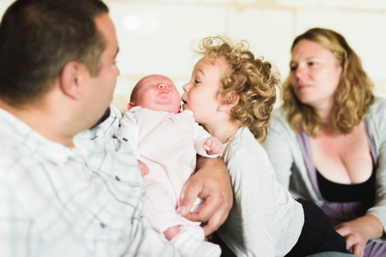 Un homme tient un bébé dans ses bras tandis qu'un jeune enfant aux cheveux bouclés se penche pour l'embrasser sur la joue. Une femme aux cheveux blonds est assise à côté d'eux, souriant et regardant les enfants. La famille semble être assise ensemble dans un cadre détendu et intime, probablement leur maison, incarnant une naissance sereine.