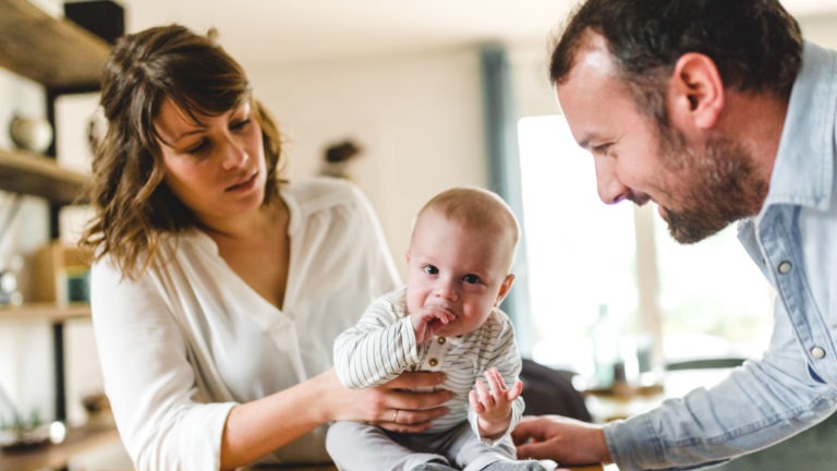 Une femme et un homme sont assis à l'intérieur, souriant à un bébé assis sur une table entre eux. Le bébé porte une combinaison rayée et a une main dans la bouche. Des étagères et des objets de décoration sont à l'arrière-plan, éclairés par la lumière naturelle. Tous semblent heureux et engagés avec le bébé, chérissant l'importance des souvenirs d'enfance.