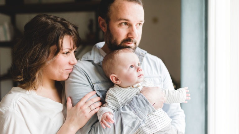 Un couple se tient près d'une fenêtre, tenant un bébé dans ses bras. La femme, en chemise blanche, regarde dehors avec un léger sourire. L'homme, en chemise en jean clair, berce le bébé qui regarde également dehors. La lumière naturelle illumine doucement la scène, capturant l'importance des souvenirs d'enfance en cours de fabrication.