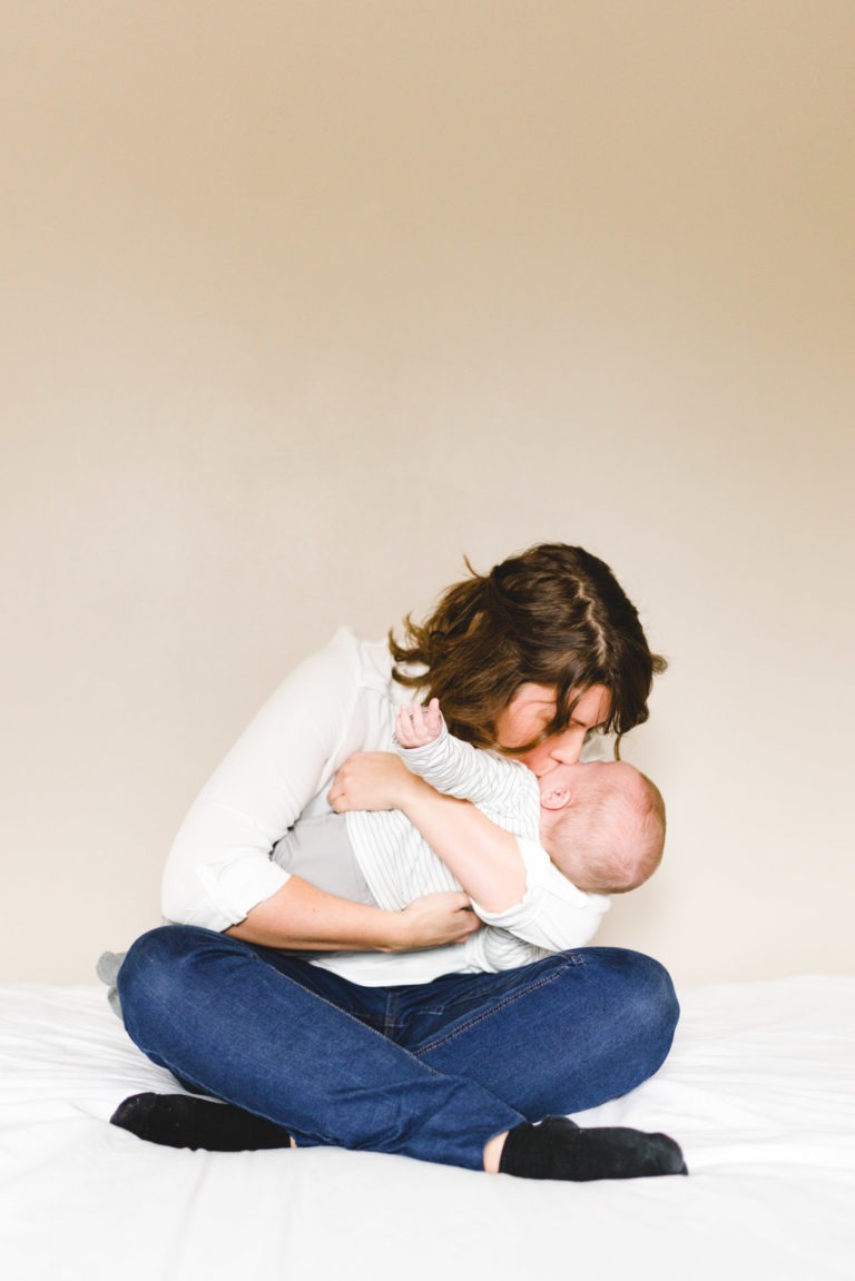 Une femme aux cheveux bruns courts est assise en tailleur sur un lit blanc, vêtue d'une chemise blanche et d'un jean bleu. Elle tient et embrasse tendrement un bébé vêtu d'une tenue rayée blanche et grise, capturant l'importance des souvenirs d'enfance sur fond d'un mur beige uni.