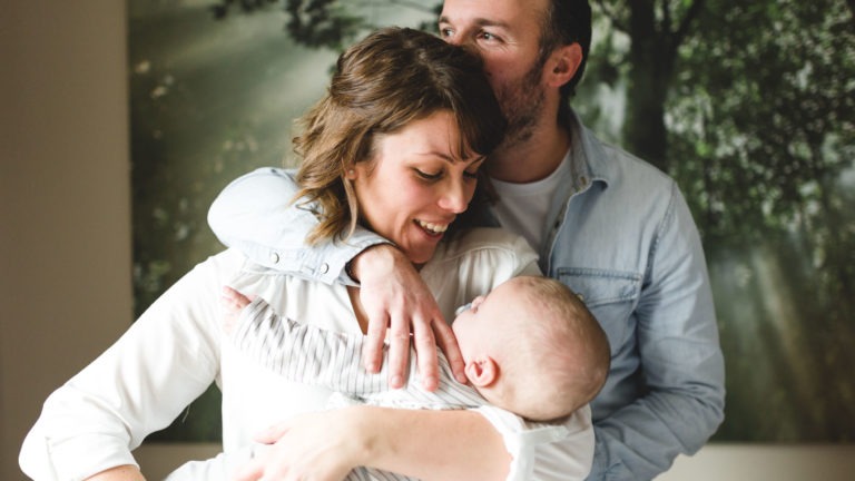 Une femme souriante tient un bébé vêtu d'une combinaison rayée, tandis qu'un homme, debout derrière elle, les enlace doucement. Il a les yeux fermés, posant son front sur la tête de la femme. Ils se trouvent dans une pièce chaleureusement éclairée avec un décor sur le thème de la nature, avec des arbres et la lumière du soleil filtrant à travers les feuilles - une image qui capture l'importance des souvenirs d'enfance.