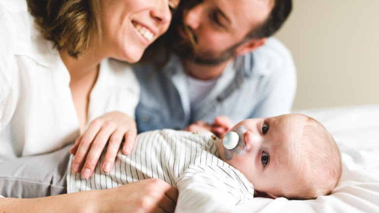 Un bébé avec une tétine est allongé sur un lit, regardant la caméra. Le bébé porte une tenue rayée blanche et grise. Derrière le bébé, une femme souriante et un homme barbu se penchent, appréciant l'importance des souvenirs d'enfance tout en partageant un moment de tendresse en famille.