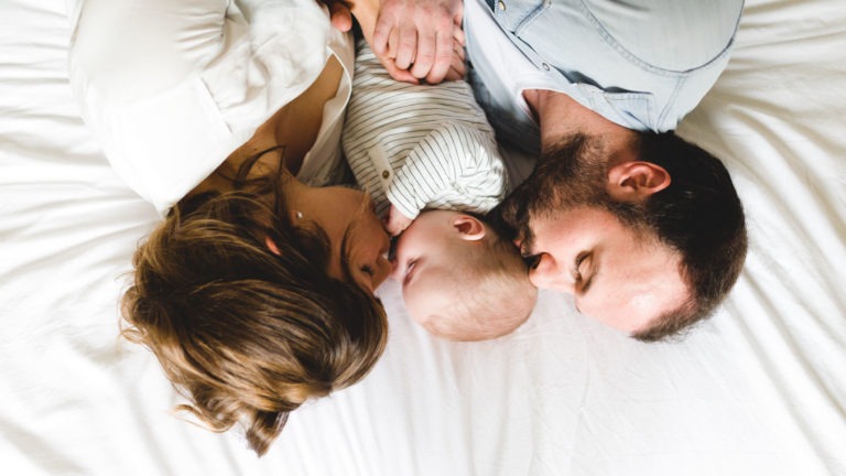 Un couple est allongé sur un lit blanc, face à face, tandis que leur bébé est couché entre eux. La femme, aux cheveux mi-longs, embrasse doucement la tête du bébé tandis que l'homme, barbu, regarde l'enfant avec amour. Leur proximité et leur tendresse soulignent l'importance des souvenirs d'enfance qui se créent dans ce moment.