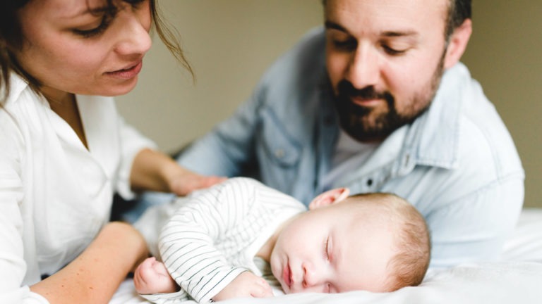 Une femme et un homme regardent avec amour un bébé qui dort paisiblement sur une couverture blanche. La femme, à gauche, place doucement son bras autour du bébé, tandis que l'homme, à droite, se penche vers lui. La scène capture magnifiquement l'importance des souvenirs d'enfance alors qu'ils chérissent ce moment tendre.