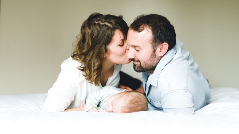 Une femme et un homme sont allongés sur un lit blanc, la femme embrassant l'homme sur la joue. Entre eux se trouve un bébé endormi vêtu d'une combinaison rayée. Tous trois semblent satisfaits et détendus, créant un moment familial chaleureux qui souligne l'importance des souvenirs d'enfance. L'arrière-plan est un mur uni aux tons neutres.