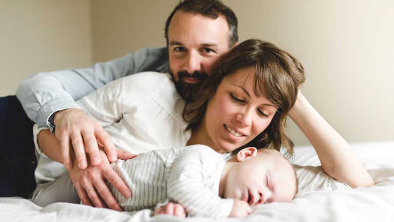 Un homme barbu et une femme aux cheveux mi-longs sont allongés sur un lit et sourient à la caméra. Devant eux, un bébé en pyjama rayé dort paisiblement sur le côté. L'homme tient le bras de la femme tandis qu'elle repose sa tête sur son autre main, capturant l'importance des souvenirs d'enfance et dégageant chaleur et lien familial.