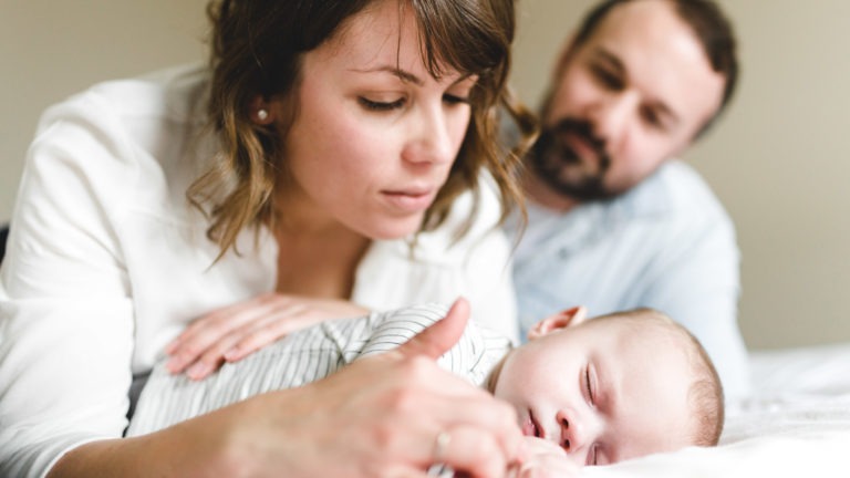 Une femme en haut blanc touche doucement la main d'un bébé endormi, tandis qu'un homme barbu regarde tendrement en arrière-plan. Le bébé est allongé sur un lit blanc dans une tenue rayée. Cette scène réconfortante respire la chaleur et le lien familial, soulignant l'importance des souvenirs d'enfance.