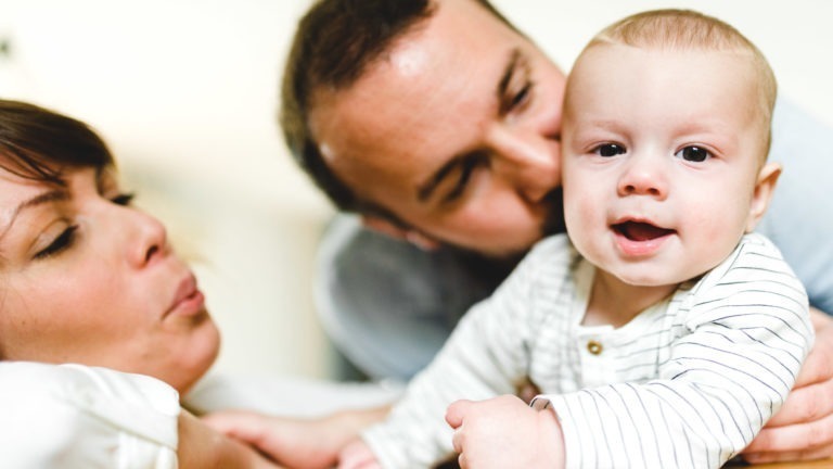 Un bébé en grenouillère rayée sourit à la caméra, allongé sur le ventre. Derrière le bébé, deux adultes le regardent affectueusement, soulignant l'importance des souvenirs d'enfance. La scène est chaleureuse et intime, suggérant un cadre familial. L'arrière-plan est flou, mettant en valeur le lien.