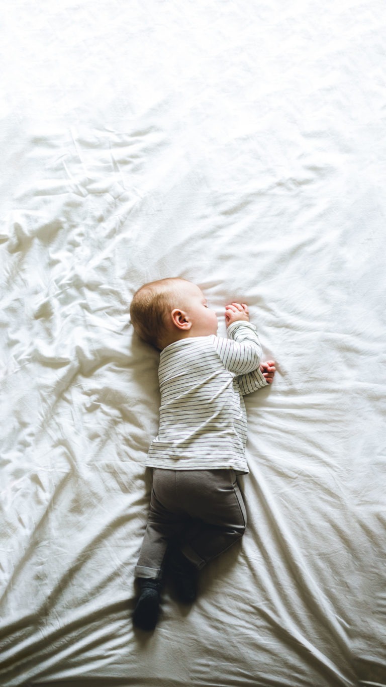 Un bébé est couché sur le côté, dormant paisiblement sur un lit aux draps blancs. Vêtu d'une chemise à manches longues gris clair et d'un pantalon gris plus foncé, le bébé aux cheveux clairs est placé au centre de l'image, entouré de draps froissés, capturant l'importance sereine des souvenirs d'enfance.