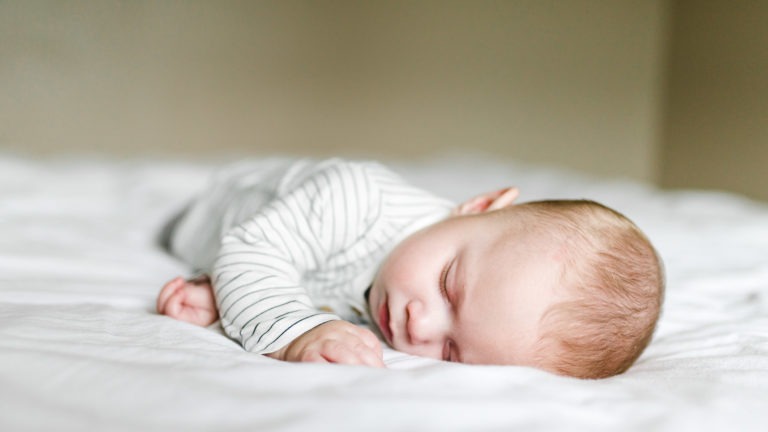 Un bébé vêtu d'une combinaison blanche à manches longues avec de fines rayures grises dort sur un lit aux draps blancs. Le bébé est allongé sur le ventre, la tête tournée sur le côté et un bras replié en dessous, incarnant l'importance sereine des souvenirs d'enfance dans un arrière-plan flou aux tons neutres.