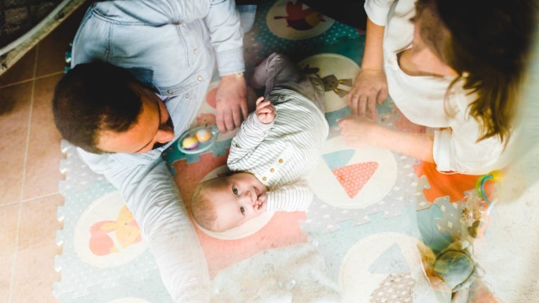 Un bébé est allongé sur un tapis coloré, le regard levé et souriant. Ils sont entourés d'un homme et d'une femme adultes qui interagissent avec le bébé, créant une atmosphère chaleureuse et ludique. Le tapis présente divers motifs ludiques, notamment un champignon et des oiseaux. Ces moments soulignent l'importance des souvenirs d'enfance pour toutes les personnes concernées.