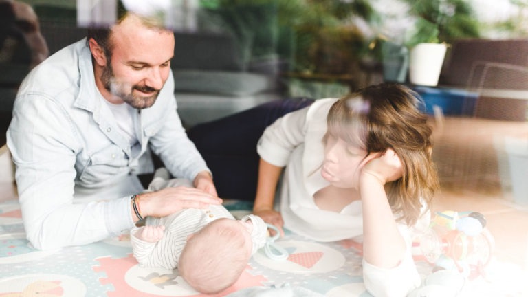 Un homme et une femme sont allongés sur un tapis de jeu coloré, en train de s'occuper d'un bébé couché sur le dos. L'homme sourit et touche doucement le bébé tandis que la femme, posant sa tête sur sa main, l'observe attentivement. Des jouets sont éparpillés dans leur espace de vie douillet, soulignant l'importance des souvenirs d'enfance.