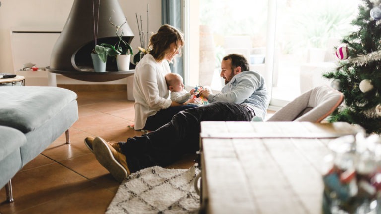 Une femme et un homme jouent avec un bébé dans un salon chaleureux doté d'une cheminée moderne, d'une plante en pot, d'un tapis et d'un sapin de Noël. La femme, assise, tient le bébé tandis que l'homme est allongé sur le sol, en train de s'occuper du nourrisson. Un canapé et une table basse sont au premier plan tandis que la lumière du soleil filtre à travers de grandes fenêtres, capturant l'importance des souvenirs d'enfance dans ce cadre chaleureux.