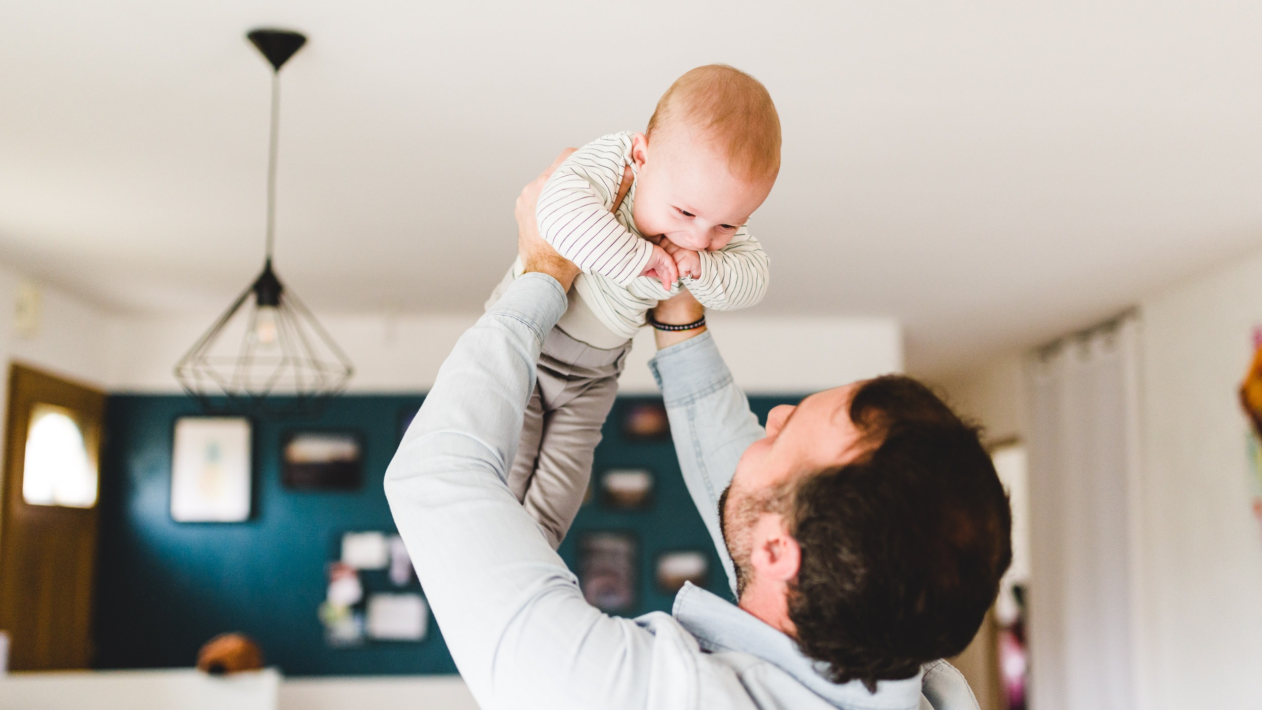 Une personne en chemise bleu clair tient un bébé souriant dans les airs. Le bébé, vêtu d'une tenue rayée blanche et grise, rit de joie tandis que la personne le regarde. Dans un salon moderne aux murs blancs, au sol sombre et à un luminaire minimaliste, ils capturent l'importance des souvenirs d'enfance.