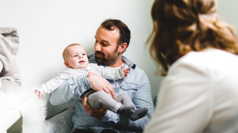 Un homme tient un bébé vêtu d'une tenue rayée blanche et grise, assis sur un lit. Le bébé regarde curieusement sur le côté. L'homme, vêtu d'une chemise bleu clair, sourit au bébé. Une femme aux cheveux longs châtain clair, partiellement visible de derrière, est assise à proximité, interagissant avec eux - une scène soulignant l'importance des souvenirs d'enfance.