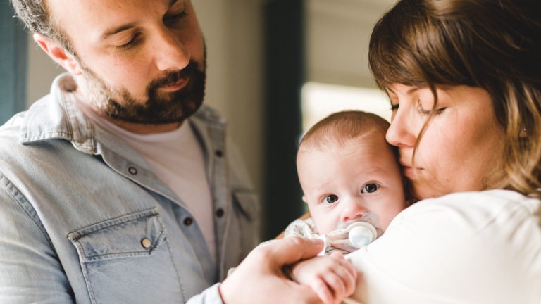 Un homme barbu et une femme câlinent un bébé. Le bébé, tenu étroitement par la femme qui semble lui embrasser la tête, regarde directement la caméra en tenant une tétine. L'homme se tient à proximité avec une expression douce, soulignant l'importance des souvenirs d'enfance dans leur étreinte chaleureuse.