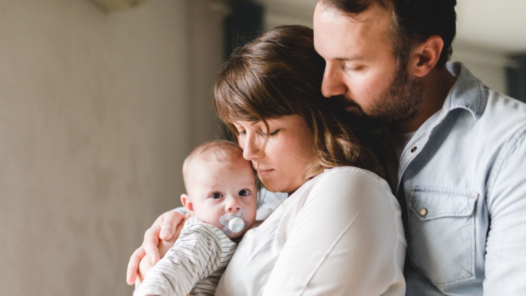 Une femme tient un bébé avec une tétine, le bébé regarde l'objectif. La femme a les yeux fermés et l'homme debout derrière elle pose son visage près du sien, pour le réconforter. Ils sont habillés de manière décontractée, reflétant la tendresse et le lien familial - une belle capture soulignant l'importance des souvenirs d'enfance.