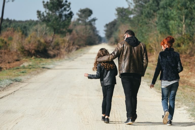 Une famille de trois personnes, qui semblent être des âmes sœurs, marche sur un chemin de terre bordé d'arbres. L'homme, vêtu d'une veste en cuir marron et d'un pantalon noir, passe son bras autour d'une fille aux cheveux longs et bouclés, portant une veste noire. Une femme aux cheveux roux, vêtue d'un gilet en fourrure noire et d'un jean, marche à côté d'eux. Des arbres et des broussailles bordent le chemin.