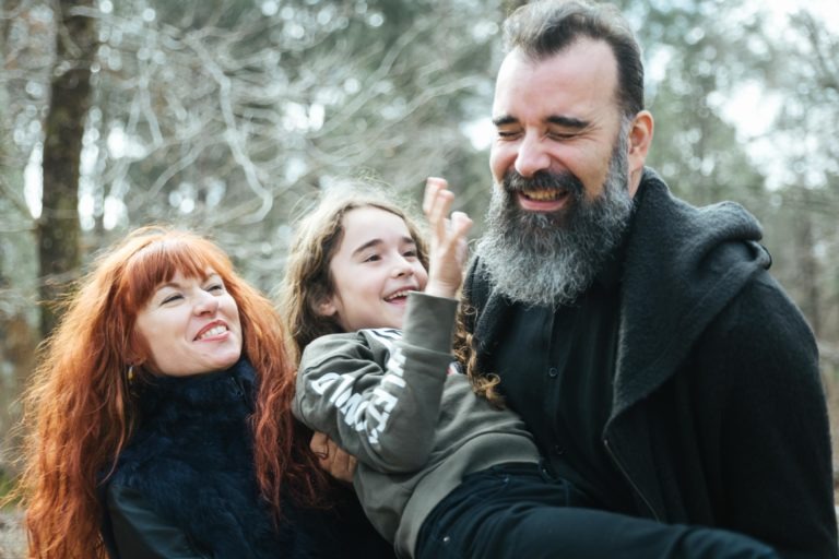 Une femme aux longs cheveux roux, vêtue de vêtements sombres, et un homme barbu en tenue noire tiennent dehors un enfant aux cheveux longs qui rit. Les âmes sœurs interagissent joyeusement dans un décor naturel aux arbres nus. Les deux adultes sourient tandis que l'enfant agite la main de manière enjouée.