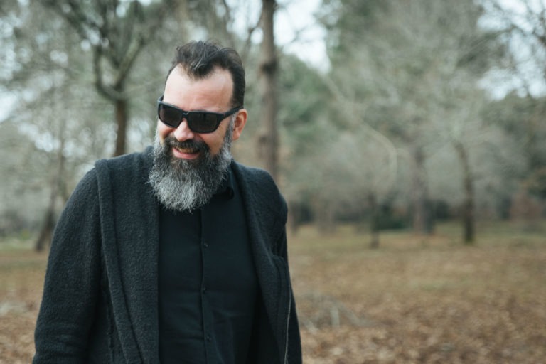 Un homme barbu aux cheveux noirs et aux lunettes de soleil sourit en se promenant dans un parc couvert de feuilles. Il porte un manteau sombre sur une chemise noire. L'arrière-plan présente des arbres nus et une atmosphère légèrement brumeuse, suggérant une journée froide d'automne ou d'hiver, parfaite pour que les âmes sœurs partagent des moments de calme ensemble.