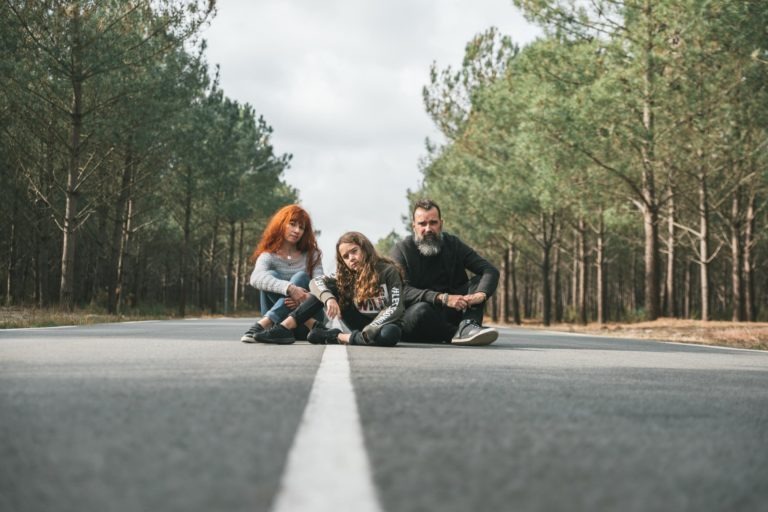 Trois personnes sont assises en tailleur au milieu d'une route pavée entourée de grands pins. Le groupe, composé d'un homme barbu et de deux jeunes femmes aux cheveux longs, est serré de manière détendue, comme des âmes sœurs, avec une seule ligne blanche qui s'étend au loin. La scène évoque une ambiance tranquille et contemplative.