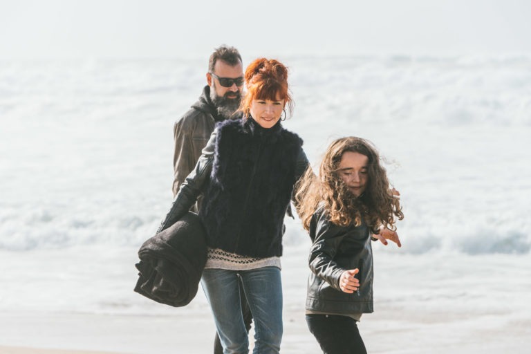 Une femme aux cheveux roux, vêtue d'un manteau de fourrure sombre, marche le long d'une plage avec son âme sœur aux lunettes de soleil et une jeune fille aux cheveux longs et bouclés. L'homme en veste marron porte une couverture, et les vagues s'écrasent en arrière-plan sous un ciel nuageux.