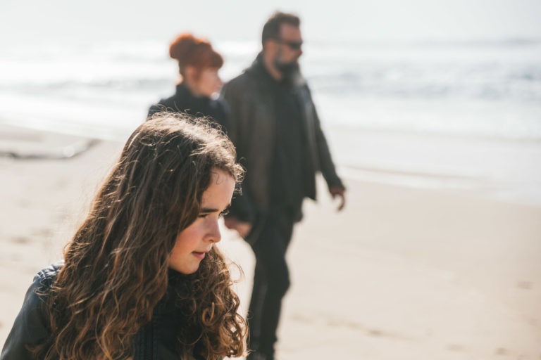 Une jeune fille aux cheveux bouclés semble pensive tandis qu'elle marche le long d'une plage. À l'arrière-plan, dans un flou, deux adultes se tiennent la main, tous deux vêtus de vêtements sombres et portant des lunettes de soleil – peut-être des âmes sœurs. Les vagues de l'océan s'échouent sur le rivage sablonneux sous un ciel clair.