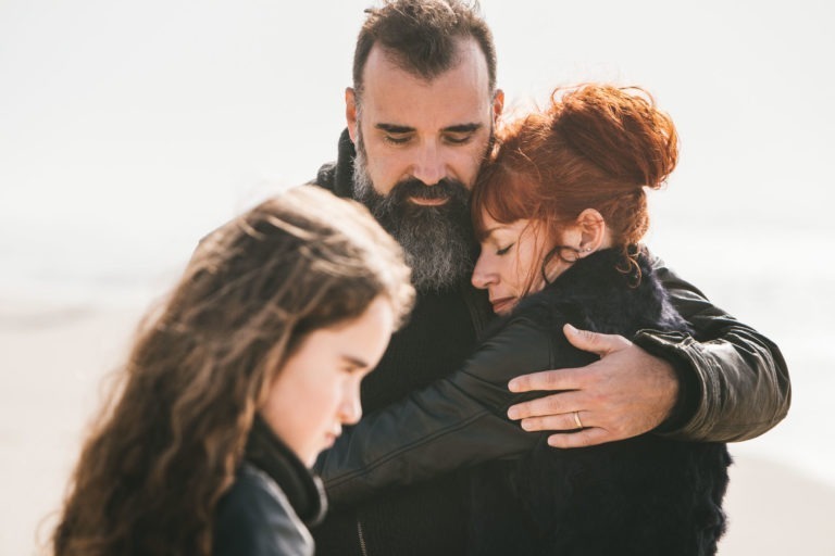 Un homme barbu enlace une femme aux cheveux roux, les yeux fermés, créant un moment serein et intime. Ils se tiennent sur une plage, âmes sœurs unies, et une jeune fille aux cheveux longs et ondulés se tient à proximité, regardant vers le bas avec une expression sombre. L'arrière-plan est flou, soulignant la connexion émotionnelle du trio.