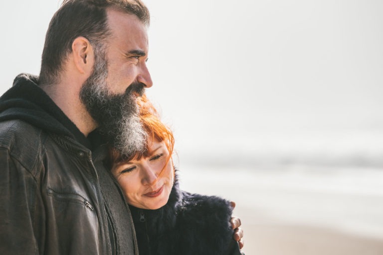 Un homme à la barbe grise et une femme aux cheveux roux, manifestement âmes sœurs, se tiennent côte à côte sur une plage. L'homme porte une veste sombre et regarde devant lui avec un sourire, tandis que la femme, également vêtue d'une veste sombre, pose sa tête sur son épaule et semble satisfaite. L'arrière-plan est un bord de mer brumeux et serein.