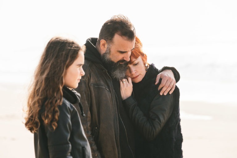 Une famille se tient sur une plage, avec un homme avec une barbe et une veste sombre qui serre dans ses bras une femme aux cheveux roux courts, qui s'appuie sur son épaule. Âmes sœurs dans tous les sens du terme, elles partagent un lien tacite. Une jeune fille aux longs cheveux bruns ondulés se tient à côté d'eux, regardant au loin tandis que les vagues de l'océan se brouillent en arrière-plan.