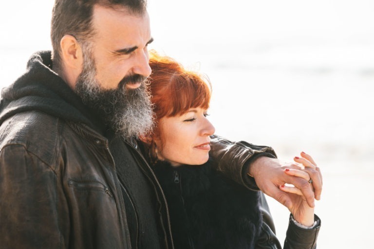 Un homme barbu portant une veste en cuir marron et une femme aux cheveux roux portant un manteau noir se tiennent côte à côte à l'extérieur. La femme touche doucement la main de l'homme, montrant une bague. Ils semblent tous deux calmes et satisfaits, comme de véritables âmes sœurs sur le fond flou et lumineux, suggérant qu'ils sont près de la mer ou de l'océan.