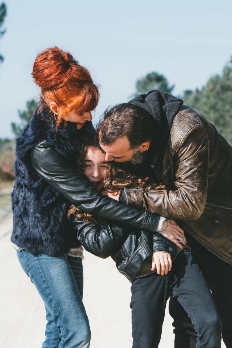 Une femme aux cheveux roux, vêtue d'un gilet noir et d'un jean, et sa sœur barbue, vêtue d'une veste en cuir marron, serrent dans leurs bras un enfant souriant, vêtu d'un sweat à capuche gris et d'un pantalon noir. Elles sont dehors par une journée ensoleillée avec des arbres et un ciel flous en arrière-plan. Le groupe semble joyeux et affectueux.