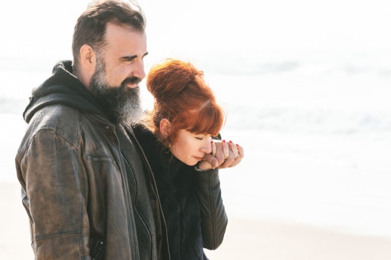 Un homme barbu, vêtu d'une veste en cuir marron et d'un sweat à capuche noir, se tient sur une plage avec la mer en arrière-plan. Une femme aux cheveux roux relevés en chignon, vêtue d'un manteau noir, se tient à côté de lui, tenant sa main sur son visage. En tant qu'âmes sœurs, elles semblent contemplatives tandis qu'elles regardent au loin.