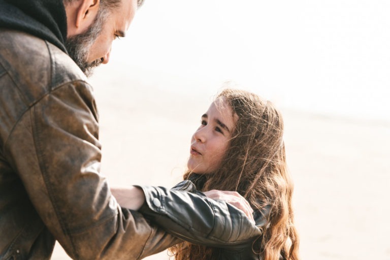 Un homme barbu, vêtu d'une veste en cuir marron, se penche et parle à une jeune fille aux cheveux longs et ondulés. La jeune fille regarde l'homme, les mains posées sur ses bras. Ils semblent être dehors, sur un fond flou et lumineux ; ils semblent être des âmes sœurs partageant un moment de tendresse.