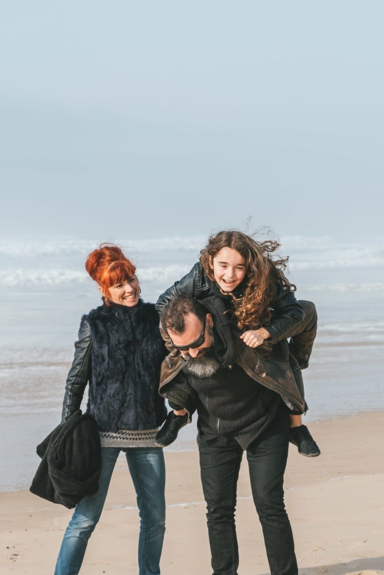 Un homme porte sur son dos un enfant souriant aux cheveux longs et bouclés sur une plage. Une femme aux cheveux roux portant un gilet de fourrure noire et tenant un manteau noir marche à côté d'eux, ressemblant à des âmes sœurs. Elles sont toutes vêtues de vêtements d'hiver avec un fond d'océan gris et des vagues visibles en arrière-plan.