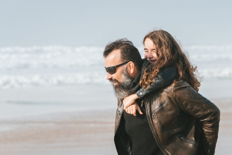 Un homme barbu, portant des lunettes de soleil et une veste en cuir marron, porte une jeune fille aux cheveux longs et bouclés sur le dos sur une plage. La jeune fille, riant joyeusement comme le font les âmes sœurs, a les bras enroulés autour des épaules de l'homme. L'océan et les vagues sont visibles en arrière-plan.