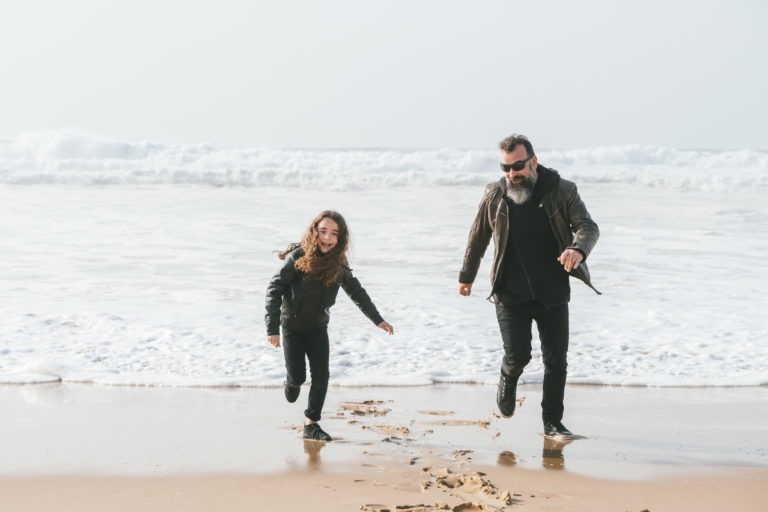 Un homme et une jeune fille, peut-être des âmes sœurs, courent sur une plage de sable avec les vagues de l'océan qui s'écrasent en arrière-plan. Tous deux sont vêtus de vêtements sombres et semblent s'amuser. L'homme a une barbe et porte des lunettes de soleil. Leurs empreintes de pas sont visibles dans le sable mouillé sous un ciel couvert.